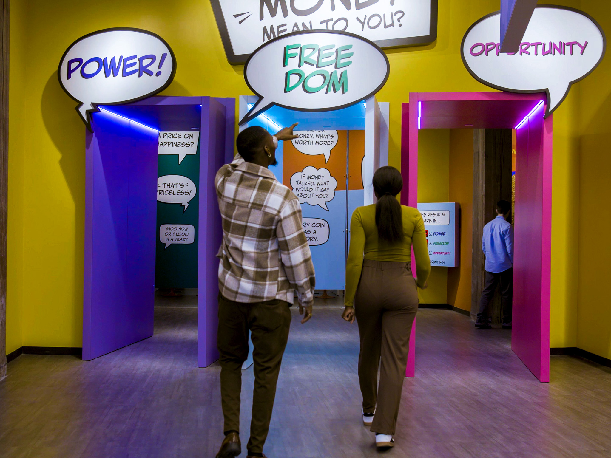 Couple walks through MoFun's bright corridor past a comic-style speech-bubble exhibit asking what money means.