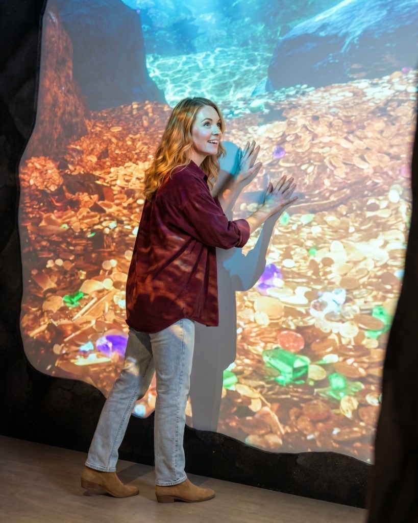 Woman presses her hands against MoFun's glowing wall of gold coins and gems at the Hoard of Gold exhibit, smiling in wonder.